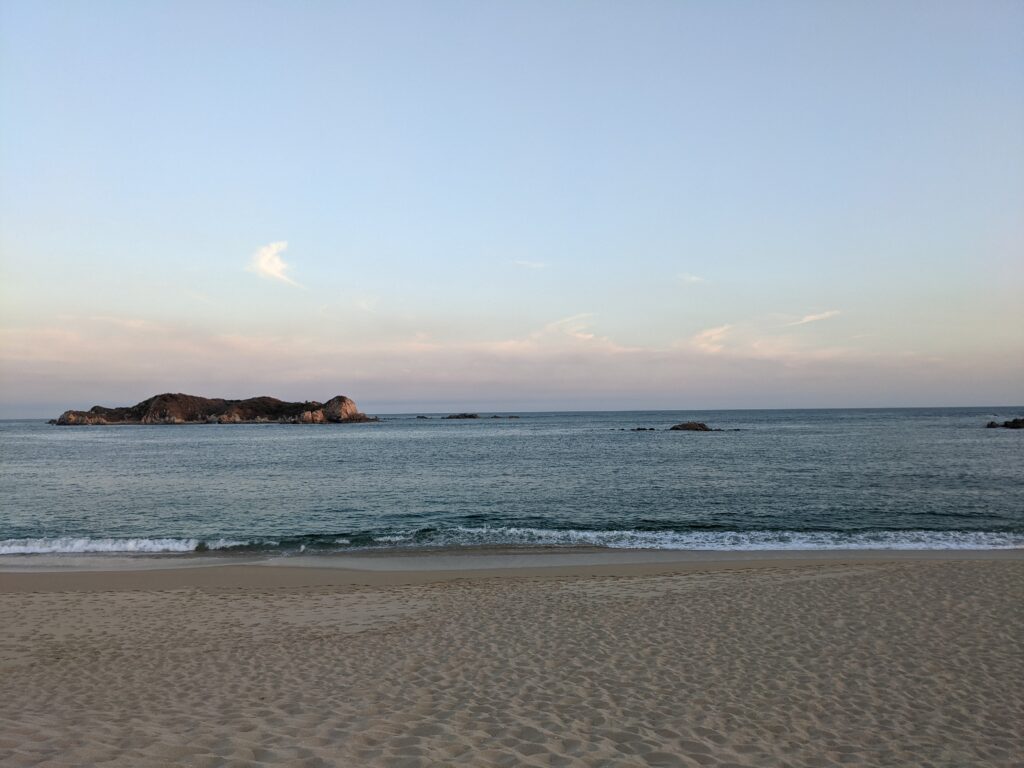 Wide view of San Agustín Beach in Huatulco at sunset, with gentle waves rolling onto the sandy shore and a rocky island in the distance under a soft pastel sky. A peaceful oceanfront scene highlighting the natural beauty of this coastal destination.
