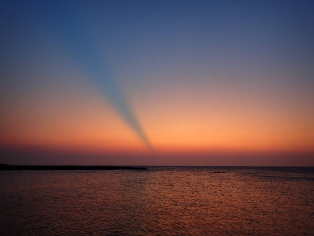 Sunset over the ocean in West Bay, Roatan, Honduras, near Turtle Beach with hues of orange, pink, and blue blending into the horizon. A shadow-like ray extends diagonally across the sky, creating a dramatic visual effect above the calm water.