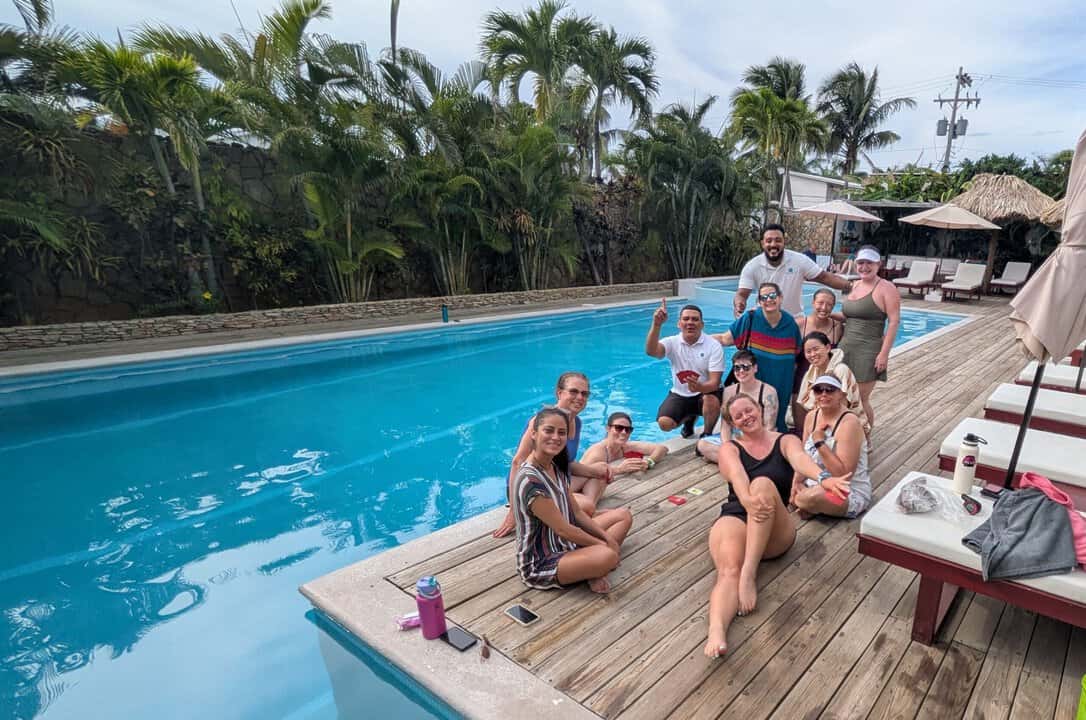 Shev and some friends play a card game, relax and laugh together beside a long blue pool surrounded by palm trees at Naboo resort. Some sit on the wooden deck while others stand nearby, creating a lively vacation atmosphere often found at roatan dive resorts. The scene highlights a casual poolside hangout with lounge chairs and lush greenery.
