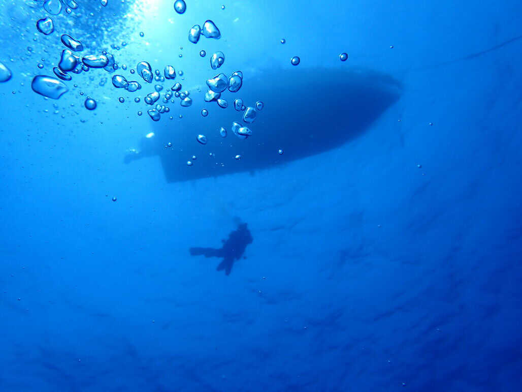 Taken from underwater looking towards the surface, the silhouette of a boat can be seen with a scuba diving drifting down beside it. Bubbles break into the frame from the photographer