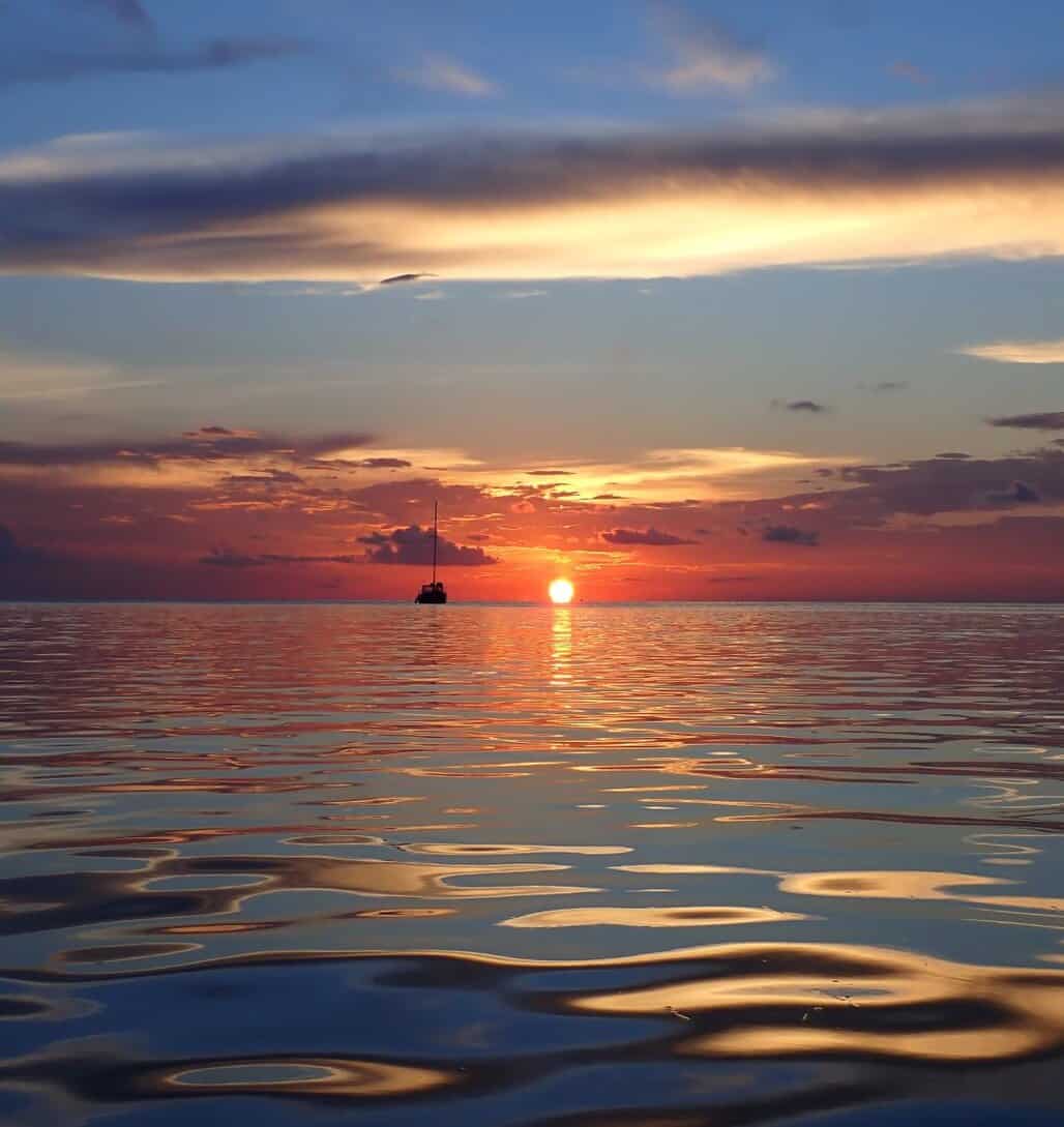 Tranquil sunset view over the ocean at one of the best Roatan dive resorts, with a lone sailboat on the horizon and warm hues reflecting off the calm water surface.
