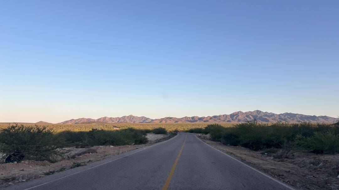 A narrow road leads straight to a mountain range. The perfect blu sky is behind, and some light green foliage scattered around. Cabo pulmo sits just beyond the mountains to the left