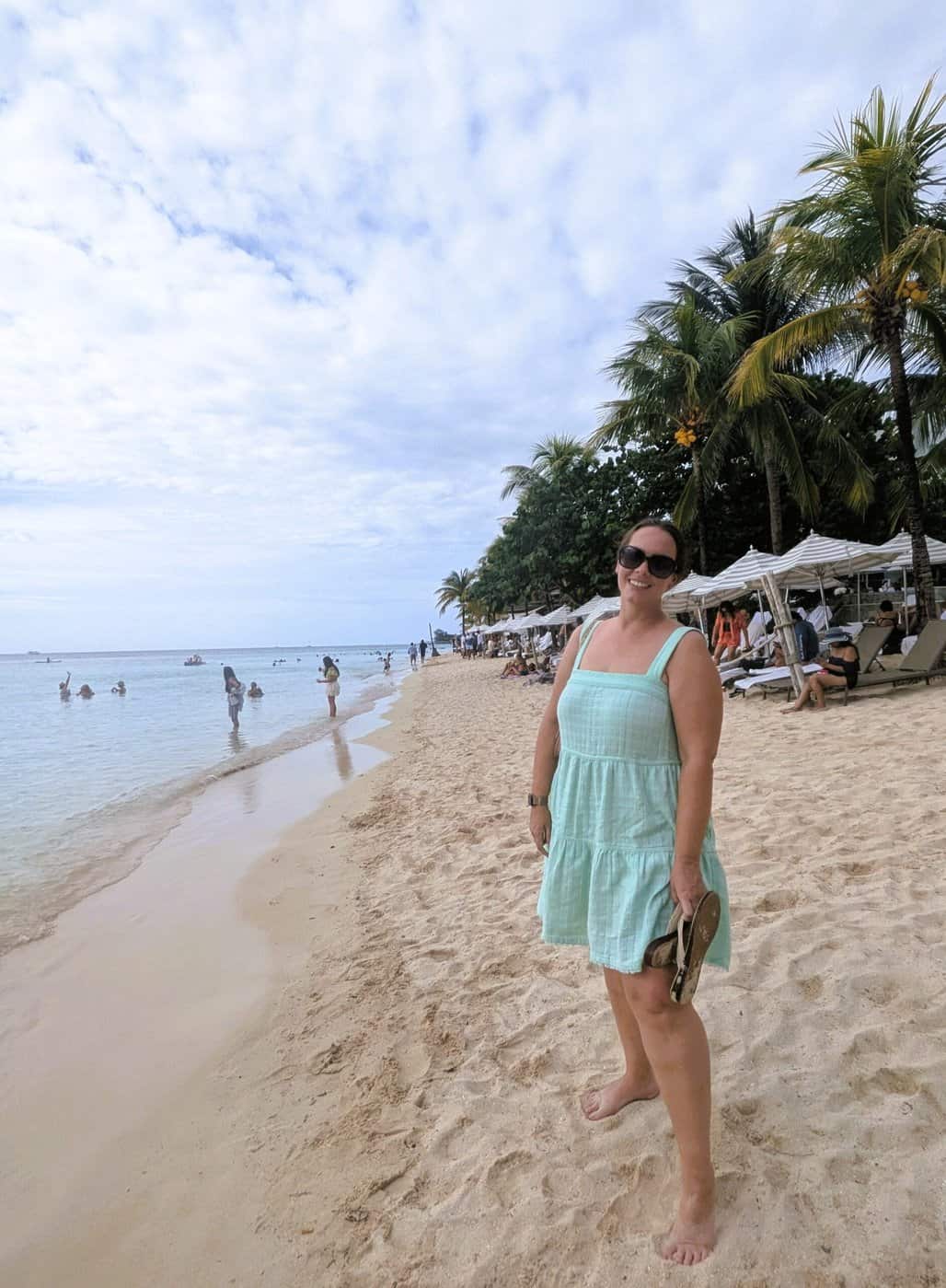 a woman stands on the white, soft sand of west bay beach in roatan. the calm caribbean water is on the left, and palm trees provide shade on the right