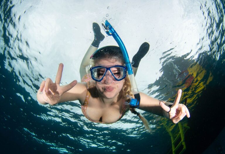 a young woman smiles at the camera through her snorkelling mask. You are looking up at her so you can see the surface of the water, and a boat and ladder just behind her.