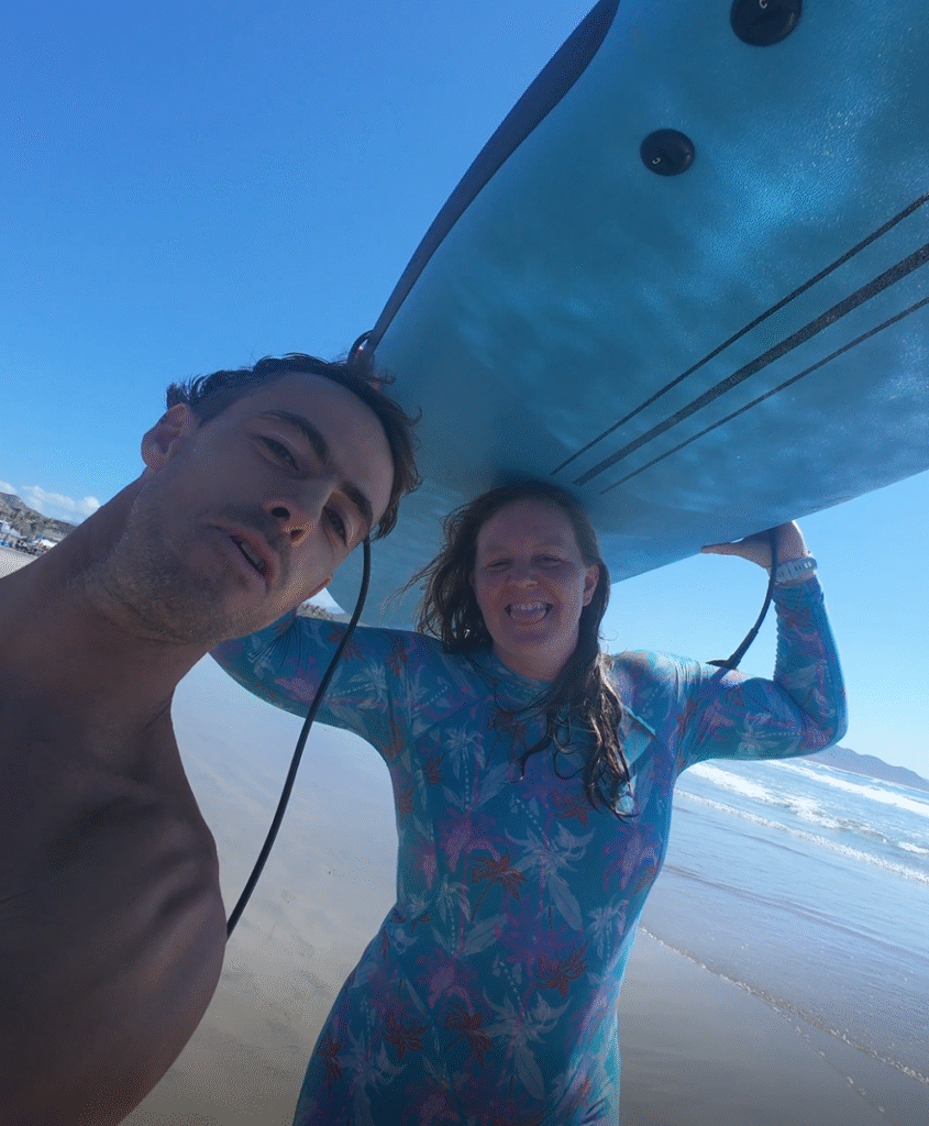 a selfie of shev, wearing  a colourful surfing suit and carrying a surfboard on her head walking to her lesson on the beach with her surfing instructo