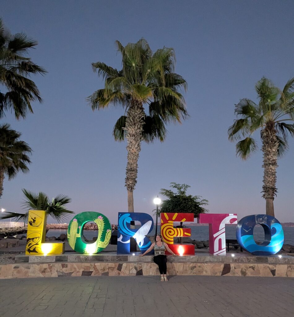 Colorful "LORETO" sign along the waterfront at dusk with palm trees behind it. A woman sits in front of the letters by the sea, marking the start of a Baja whale watching road trip in Mexico.