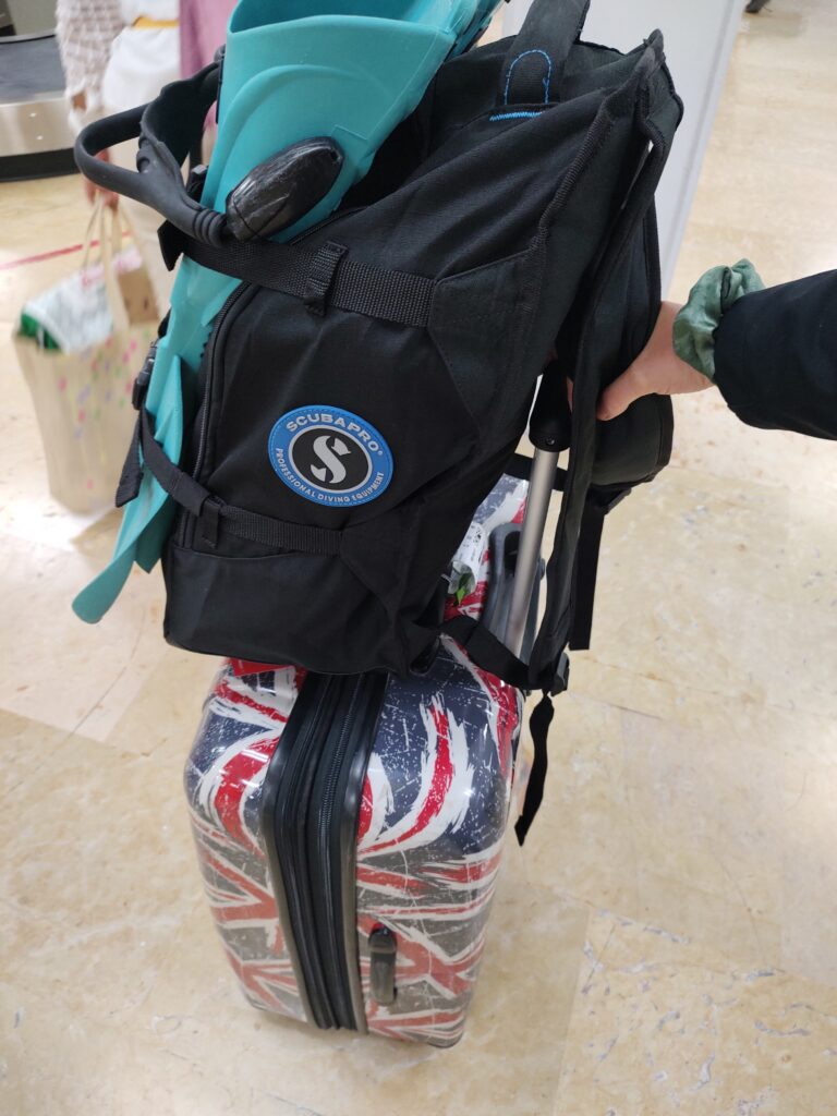 Black scuba gear backpack strapped to a rolling suitcase with turquoise dive fins tucked into the side pocket at the airport. This compact carry on dive gear setup shows a practical way of packing light and travelling with dive gear for a scuba vacation.