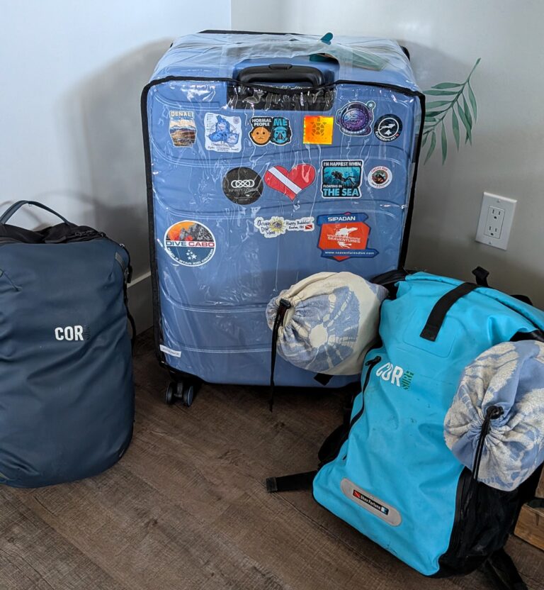 Large blue suitcase covered in dive travel stickers surrounded by waterproof bags and packed gear on a wooden floor. This setup shows an organized approach to packing dive gear and travelling with dive gear for a scuba vacation.