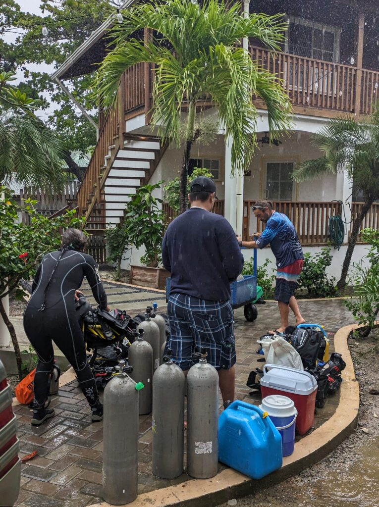 Dive group loading scuba tanks and equipment into a cart outside a dive shop during a tropical rainstorm in Roatan. Sudden heavy rain is common during parts of the year and can be a factor to consider when deciding the best time to scuba dive in Roatan.